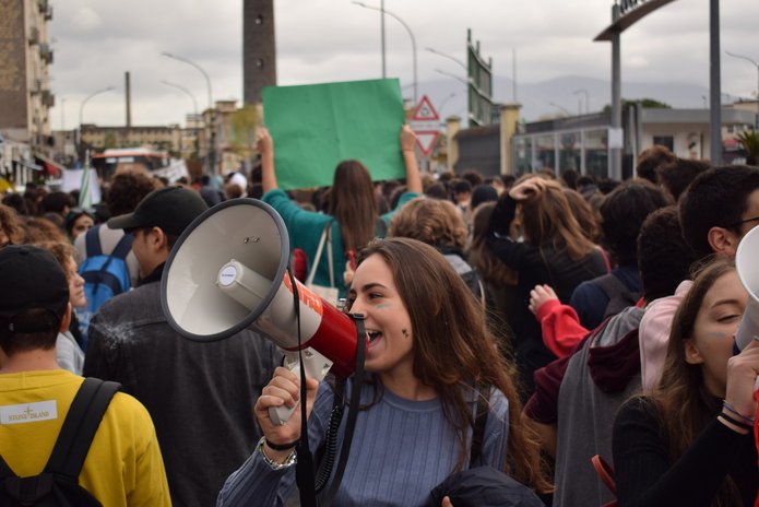 Na zdjęciu widać tłum ludzi. Na przodzie stoi dziewczyna z długimi włosami, w prawej dłoni trzyma biały megafon przez który coś krzyczy.