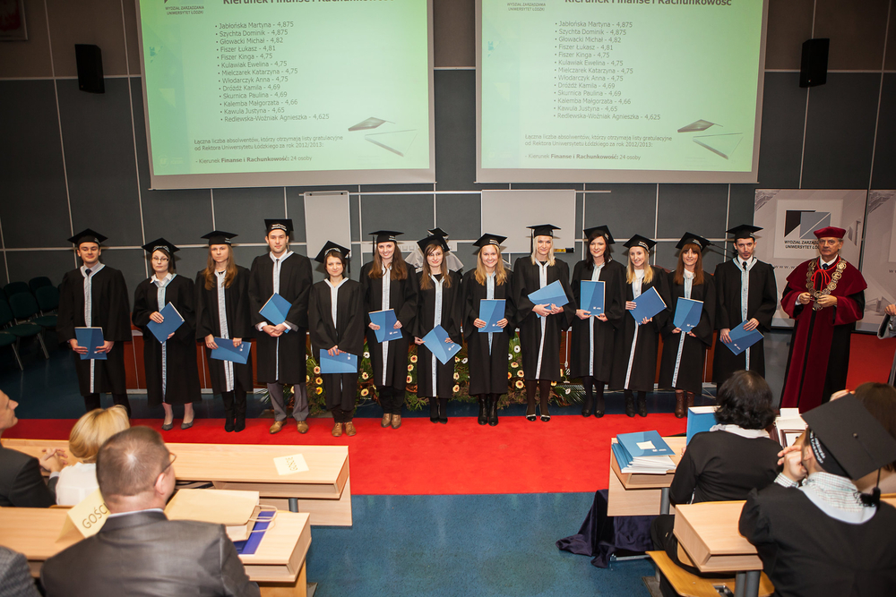 absolwenci stoją na środku auli przed prezydium, trzymają dyplomy w rękach