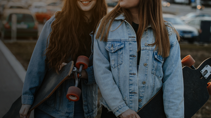 Dwie młode kobiety stoją na tle miejskiego krajobrazu z deskorolkami typu longboard w dłoniach. Obie ubrane są w jasnoniebieskie jeansowe kurtki. Jedna z nich uśmiecha się szeroko, a druga patrzy w jej stronę z lekkim uśmiechem. W tle widać zaparkowane samochody i bloki mieszkalne oświetlone ciepłym światłem zachodzącego słońca. Scena emanuje swobodą i miejskim stylem życia.