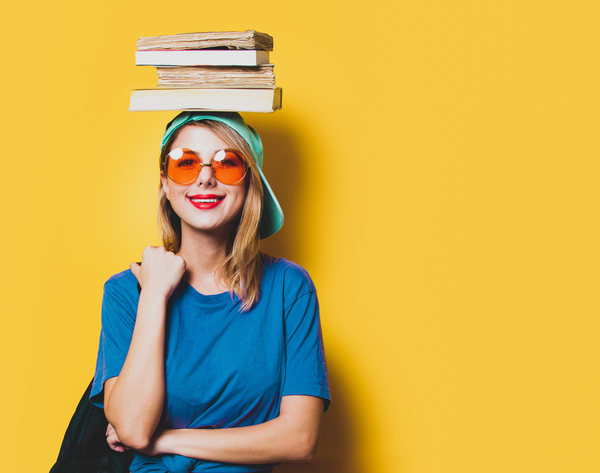 Zdjęcie przedstawia uśmiechniętą kobietę w kolorowych okularach, z książkami na głowie