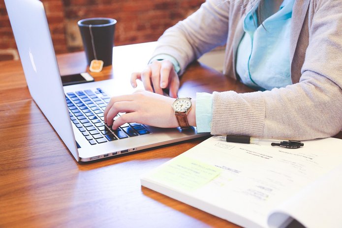 A view of a desk, with a person typing on a laptop and a notebook next to it. Their face is not visible.