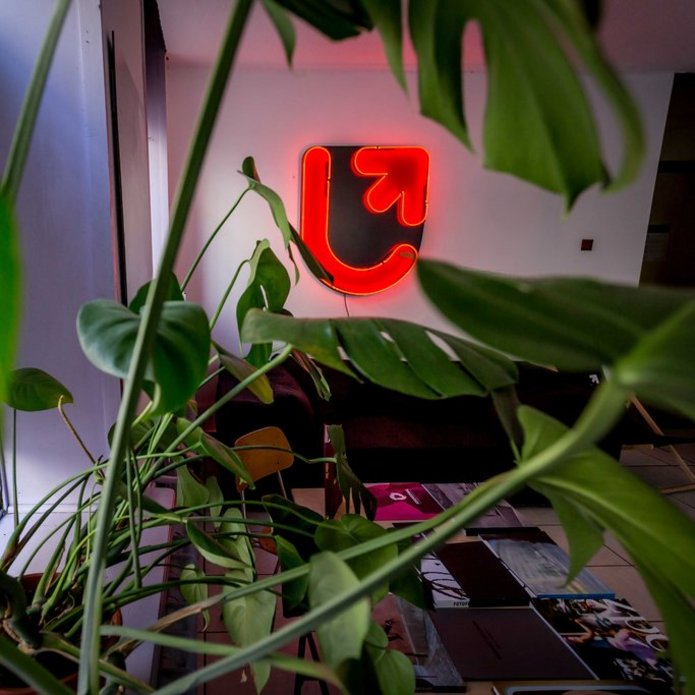A photo depicting a room with a neon sign with the UniLodz logo and potted plants