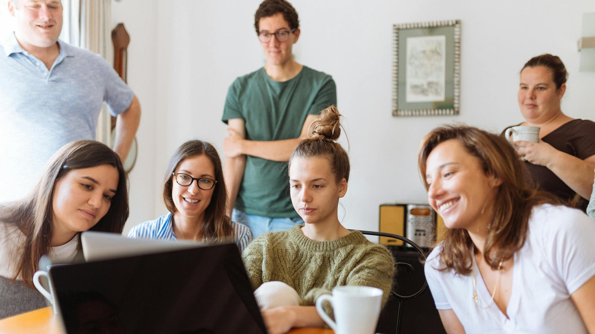 group of smiling young people sitting in a room, looking at a laptop screen