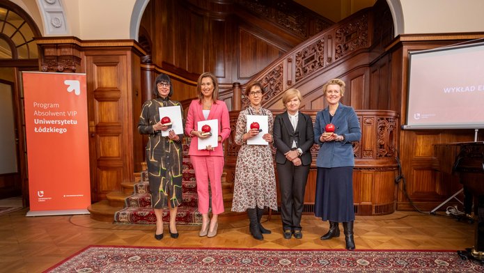 four UL VIP Alumni holding red apples of the programme with the UL Rector. They are all standing next to the stairs in the Biedermann's Palace 