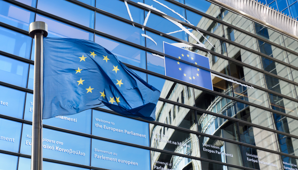 Na pierwszym planie powiewa flaga Unii Europejskiej na maszcie. W tle widoczna jest nowoczesna, przeszklona fasada budynku Parlamentu Europejskiego z napisem powitalnym w różnych językach oraz emblematem UE.